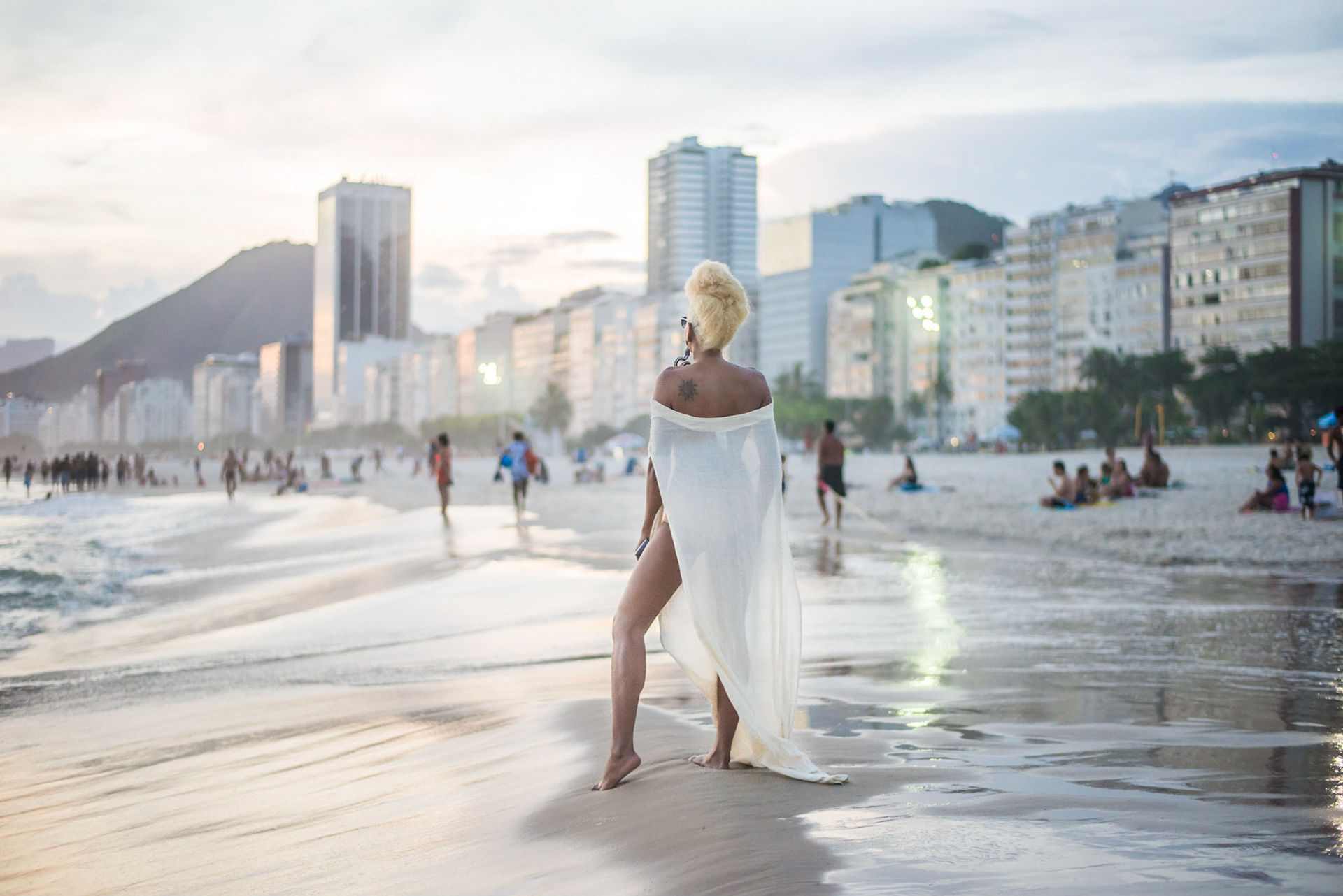 with her back to the camera, model with long white dress stares at beach. de costas para a câmera modelo de vestido longo branco olha para a praia. fashion photography, designer, clothes, dress, accessory, product, still, brand, lookbook, shooting, vibrant, colorful, beach, creative, catalogue, outfit, look.fotografia de moda, design, roupa, vestido, acessório, produto, still, marca, vibrante, colorida, praia, catálogo. rio de janeiro, copacabana