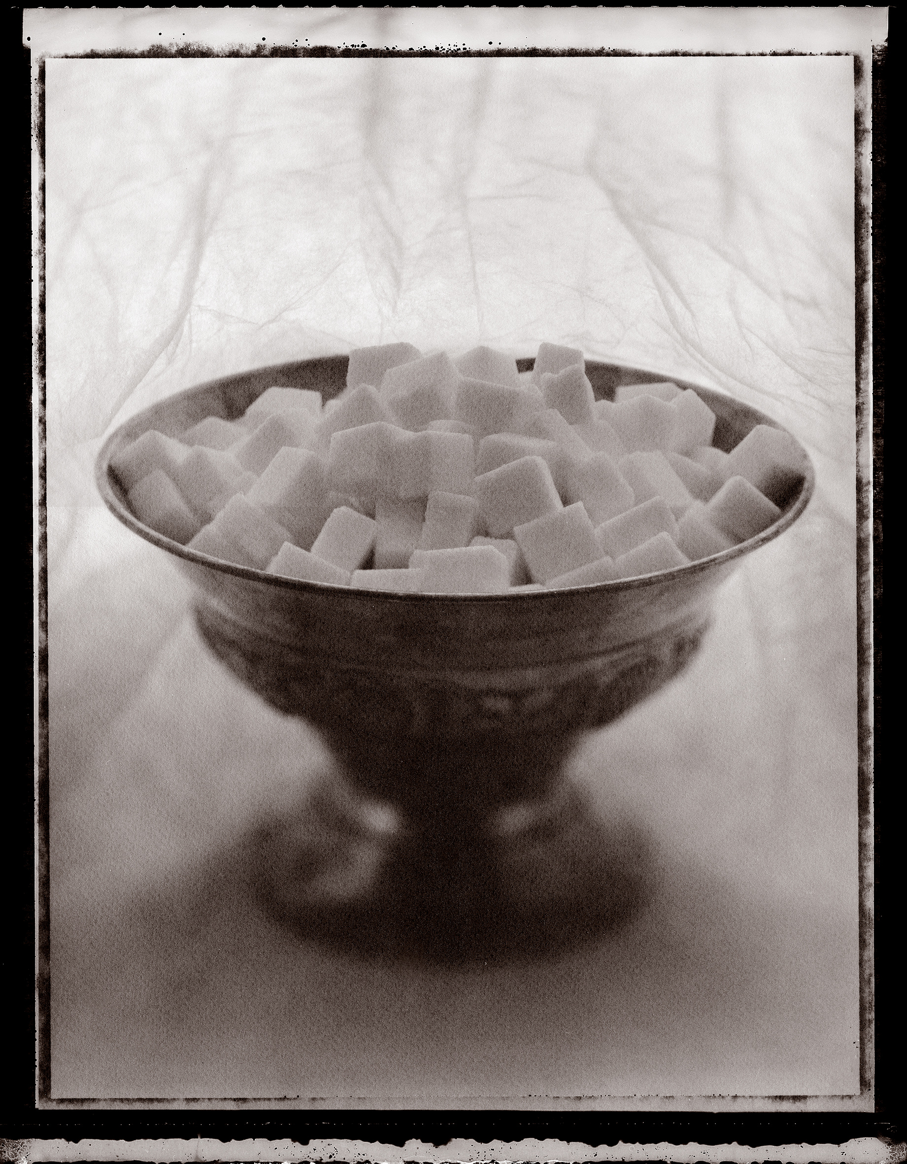 Silver bowl filled with sugar cubes against a light translucent cloth with a prominent black border. Photographed in black and white.