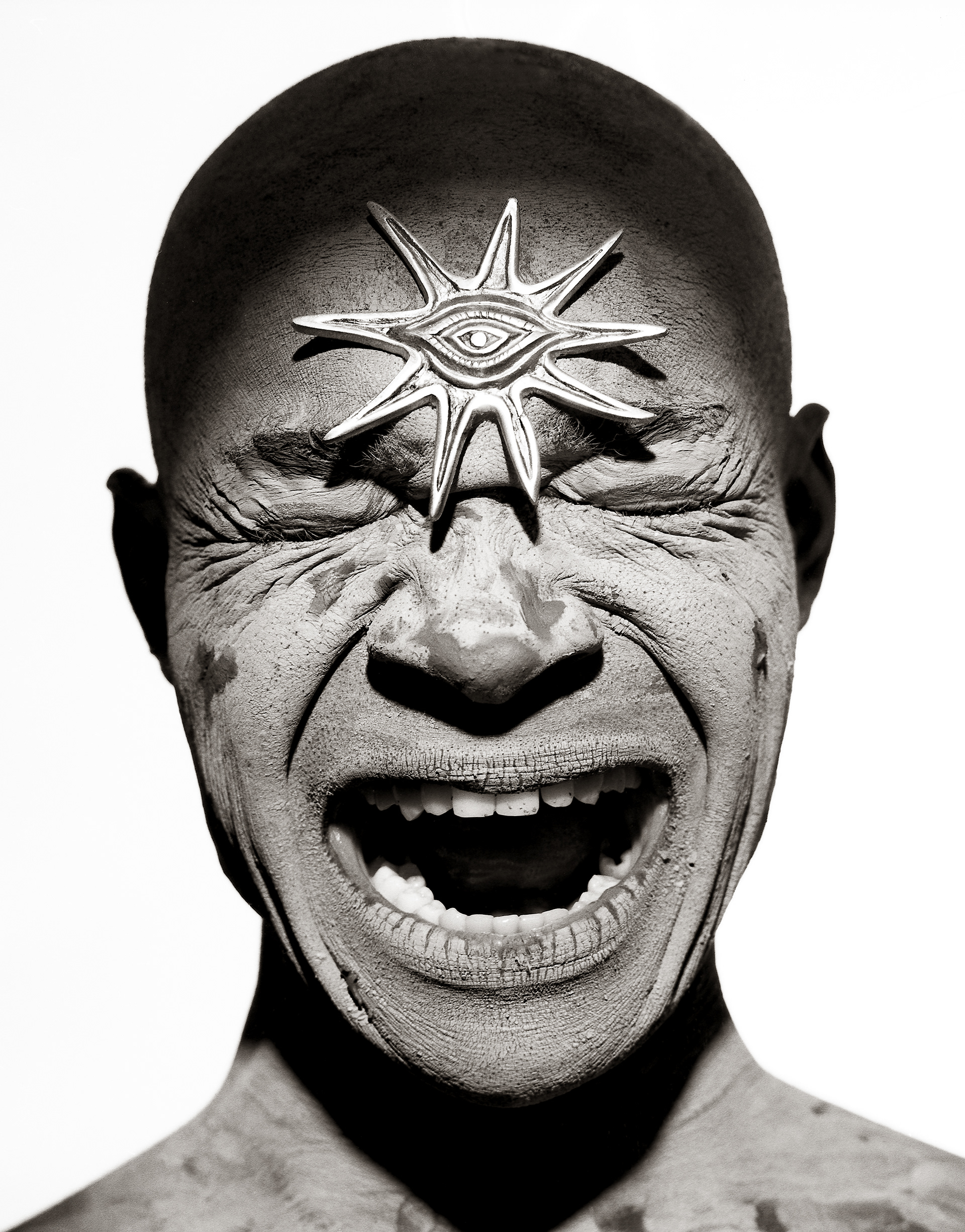 Jovans face, eyes closed, screaming,face covered in dried clay with a star shaped ornament on his forehead. Photographed against a white background in black and white