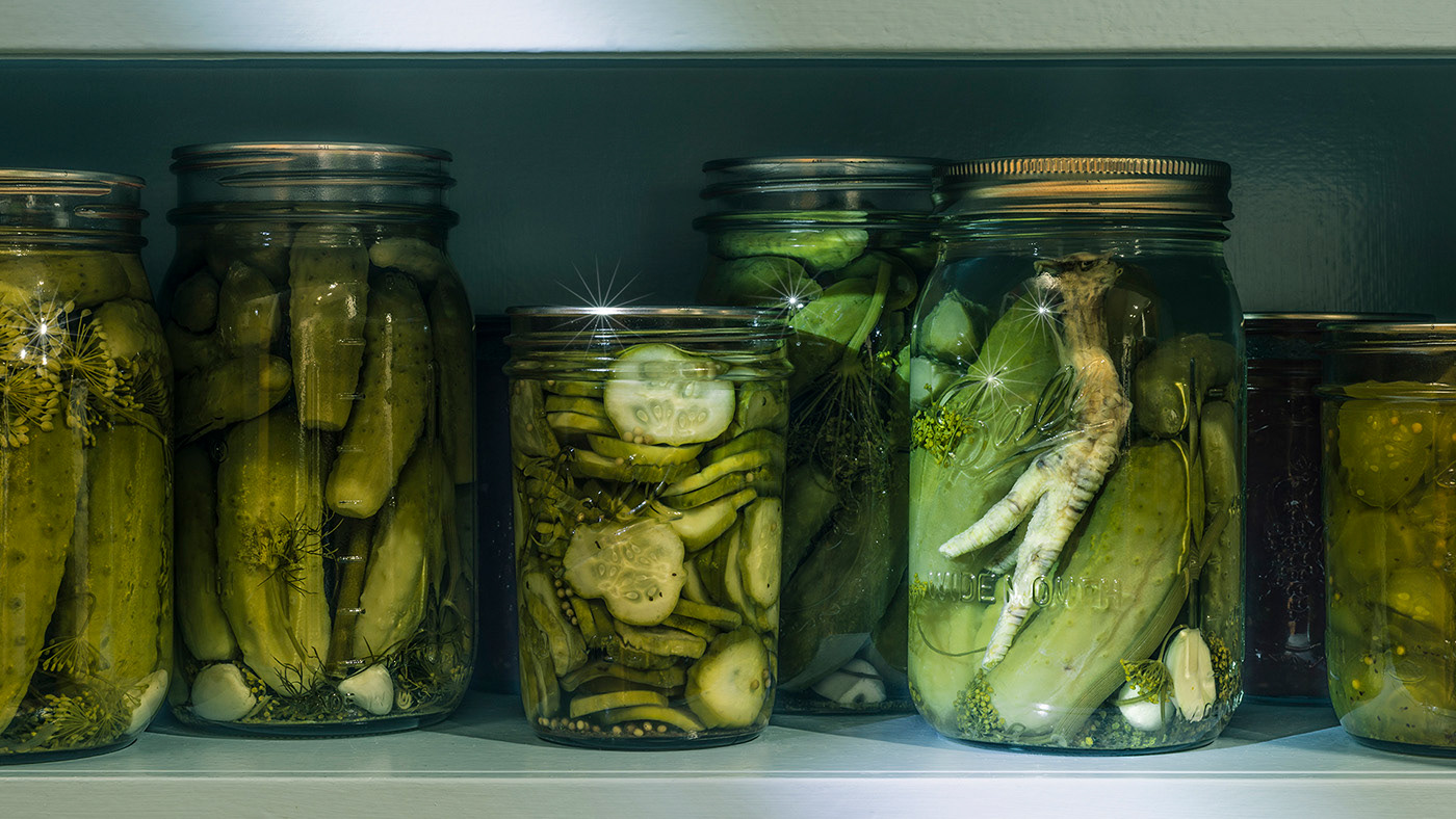 A pantry shelf filled with many jars of pickles; a chicken foot is noticed in one of the jars