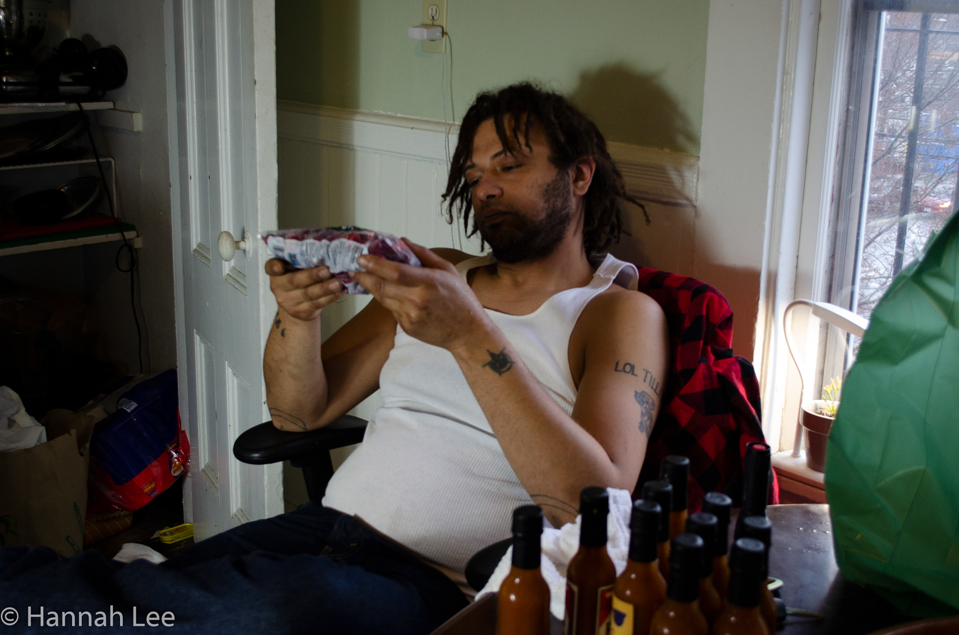 Damian inspects a bag of fresh cranberries, just purchased from Broadway market, next to a stash of hot sauce that he made from scratch and is going to sell. His friend bought the bottles and Damian grew the chilis, dried them, and let the sauce age in the fridge for a couple years. He and his friend put stickers on the hot sauce bottles last Saturday. Taken by Hannah Lee on Thanksgiving November 27, 2020 in Somerville, MA. 1/40 second f 4.5 29 mm iso 640