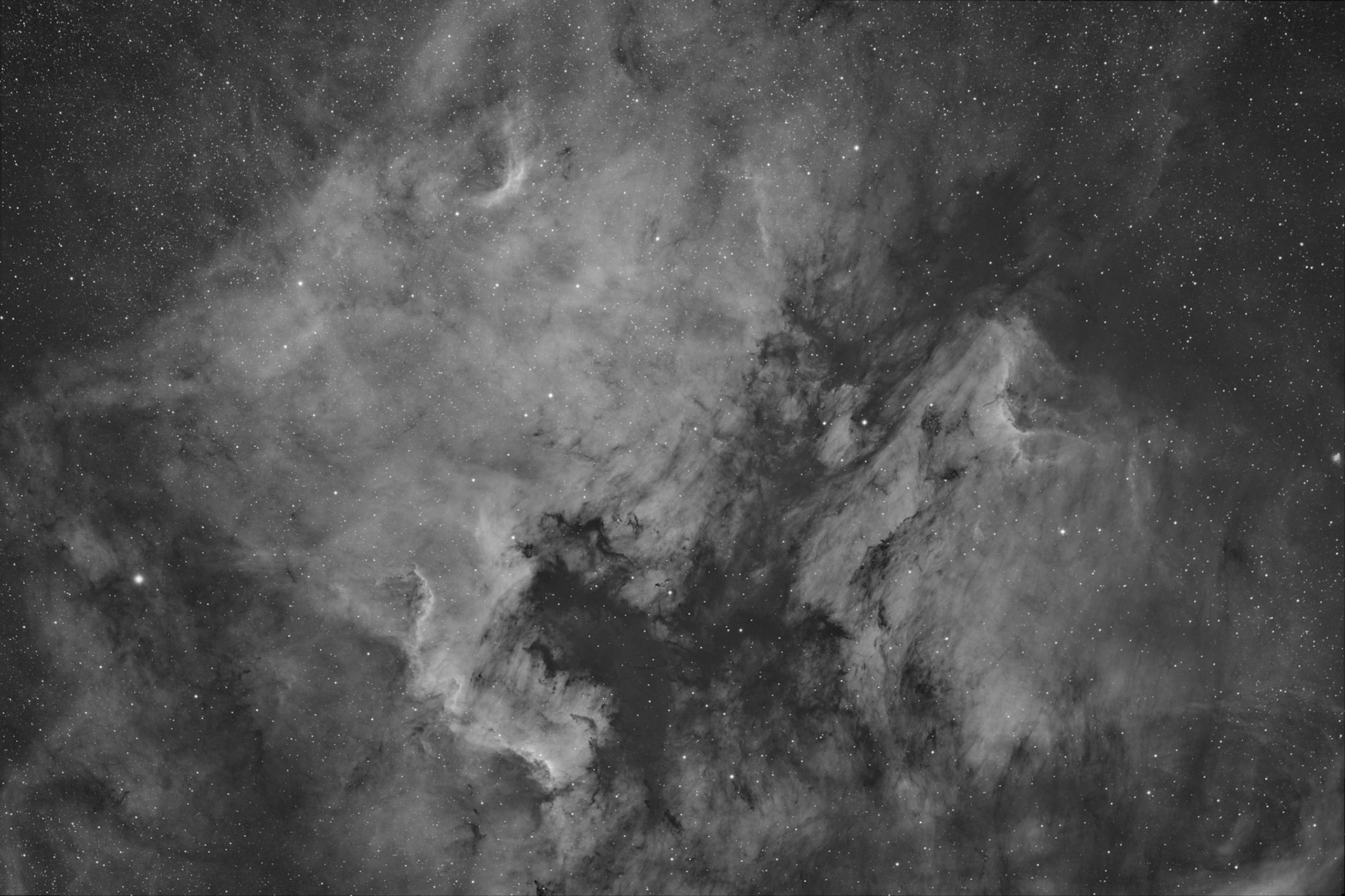North America and Pelican in Hydrogen AlphaThis one's a little different than my other astro-images. For one, you'll notice that it doesn't have any color. The other, maybe less obvious aspect, is that the nebulosity is very prominent compared to the stars in the image.  These are because I took this image with a monochrome camera through a filter that only lets through a very narrow slice of the spectrum (called a "narrow band" filter).This particular filter only let's through light that is really close to the color that Hydrogen glows at. This wavelength is towards the red end of the visual spectrum so that's why most images of nebulae tend to be red (most nebulae are primarily hydrogen).You may also notice that these particular nebulae make pretty distinct shapes. The one of the left is known as the North America Nebula and the one on the right is the Pelican Nebula.