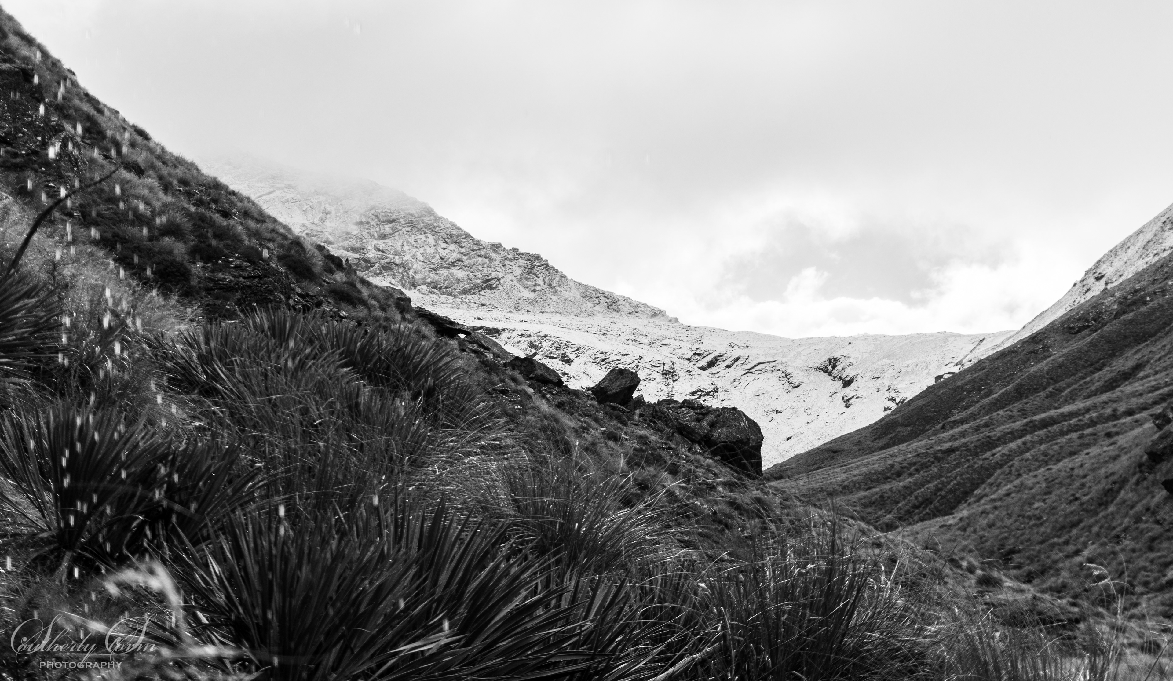 Black and while image taken from under a rock overhang, featuring heavy rain drops, spainards and distant snow covered range