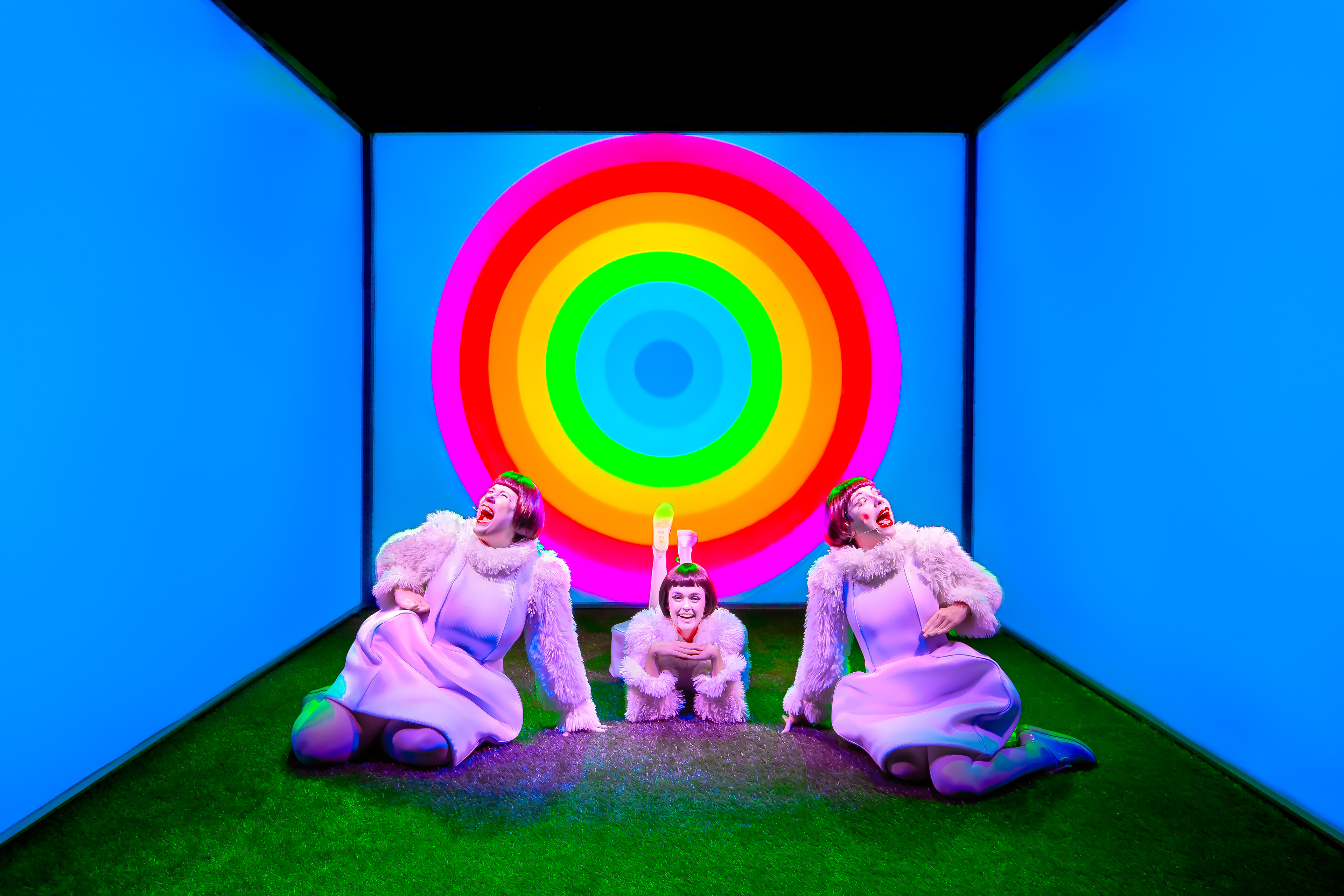 Três atrizes vestem roupas brancas de pelúcia e uma peruca escura de cabelo chanel reto. Elas são idênticas. Estão no meio de um cubo muito colorido, forrado com chão de grama artificial e iluminado por luzes também coloridas. Em perspectiva, vemos o interior do cubo, onde as atrizes estão sentadas simetricamente. Ao fundo, um arco-íris redondo ocupa a tela toda em contraste com um azul sólido que permeia as faces laterais. Elas têm sorrisos gigantescos e aparentam ser ultrafelizes.