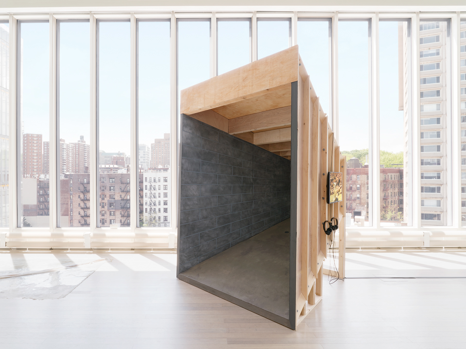 "Silver Bullet Test Site" overall shot. image description: A sculpture stands in front of a large row of windows (shades open). The sculpture is a point perspective room made out of wood, and is painted to resemble a basement on the interior. a monitor hangs on the outside of the structure.