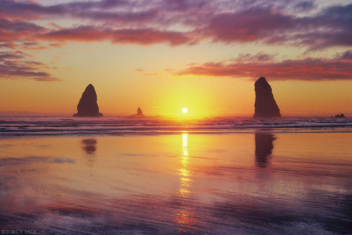 Surf rolls up beach past silhouetted sea stacks at sunset on Canon Beach, Washington