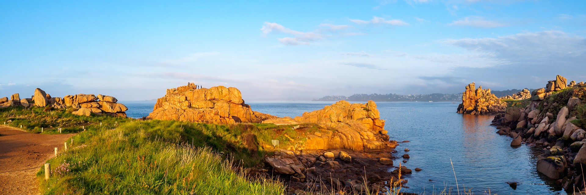 Under a blue sky during a golden sunset, a small group of daredevil divers prepared to jump off this cliff in Ploumanach, Bretagne. Wide shot with broad scenery of the pink granite, Perros-Guirec in the background.